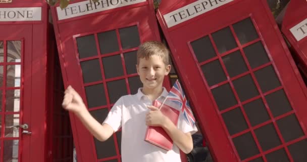 Slow motion portrait of cute boy standing outdoors alone smiling looking at camera. On the background English red telephone booths. Travelling concept. - Footage, Video
