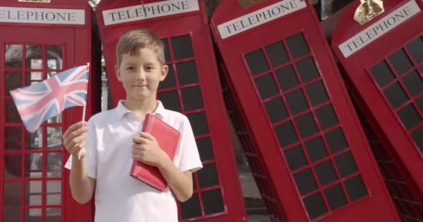 カメラを見て笑顔で屋外に立つイギリス国旗を振るかわいい少年のスローモーションポートレート。背景には英語の赤い電話ブースがあります。旅行コンセプト. - 映像、動画