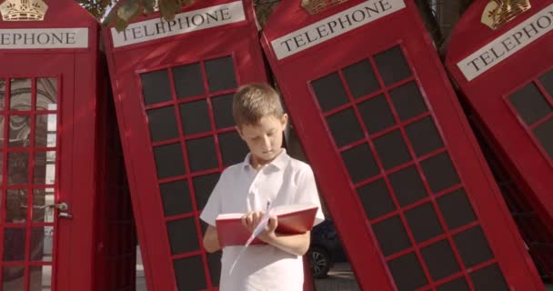 一人で屋外に立つかわいい男の子のスローモーションポートレートは、カメラを見て微笑んでいます。背景には英語の赤い電話ブースがあります。旅行コンセプト. - 映像、動画