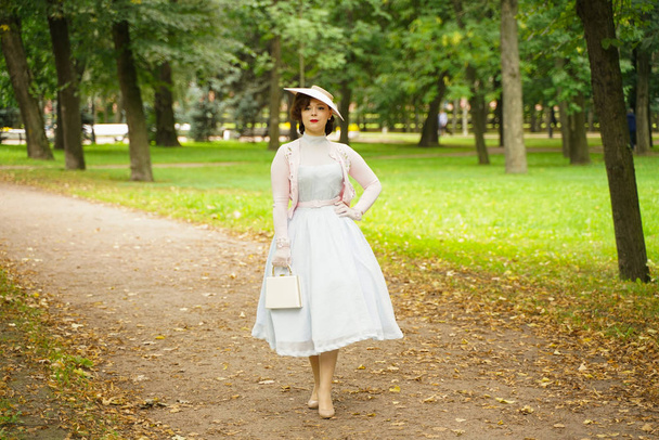 Портрет молодой женщины в винтажном платье и ретро-шляпе, гуляющей в городском парке в одиночестве
 - Фото, изображение