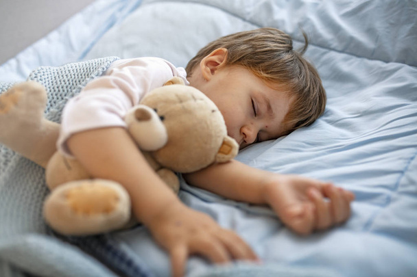 眠い頭青い枕の上で安らかに眠っている少年。赤ちゃんは彼の睡眠を必要とします!テディベアとおしゃぶりと寝ている男の子。柔らかい毛布で覆われた赤ちゃんの睡眠  - 写真・画像
