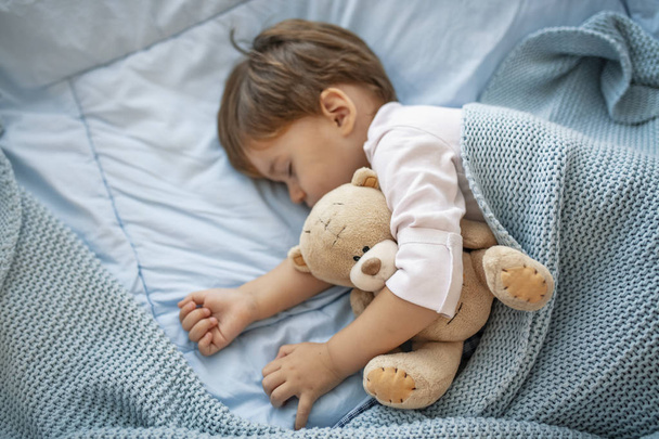 Adorable kid boy after sleeping in grey bed with toy.  Cute healthy little toddler baby boy child sleeping / taking a nap under blanket in bed while hugging teddy bear.  - Valokuva, kuva