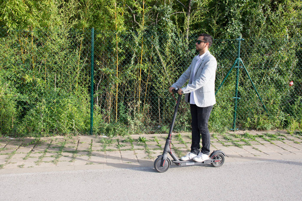 Homem adulto latino com óculos de sol, bem vestido e dirigir scooter elétrico na rua com fundo de bambu
 - Foto, Imagem