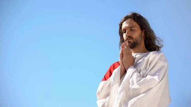 Man in robe praying to God against blue sky background, asking soul salvation - Φωτογραφία, εικόνα