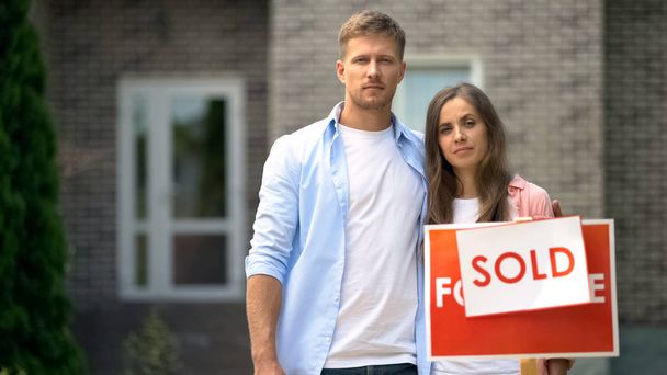 Счастливая семейная пара улыбается, стоя возле продаваемой вывески, агентства недвижимости
 - Фото, изображение