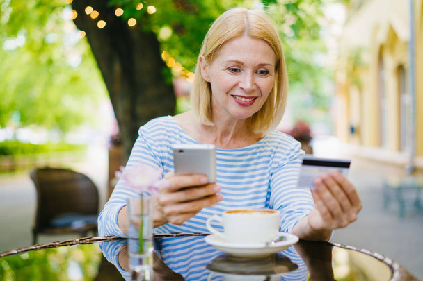 Зрелая дама платит банковской картой, используя смартфон, расслабляясь в уличном кафе.
 - Фото, изображение