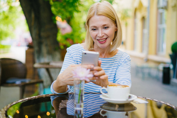 Aantrekkelijke rijpe blonde met behulp van smartphone genieten zomerdag in open lucht café - Foto, afbeelding