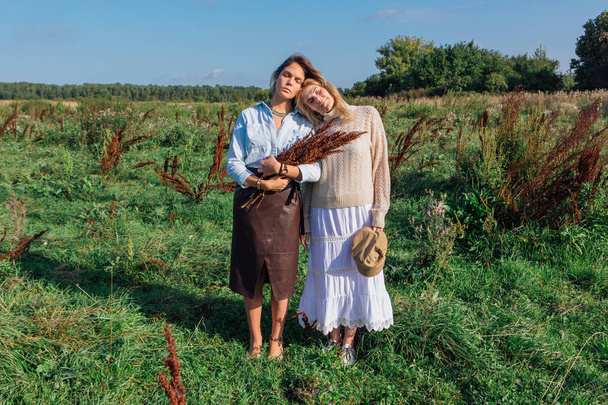 Portrait of two beautiful women in a field in late summer. Beautiful girls standing next to each other and holding bouquet made of dry brown plants. - Foto, Imagem