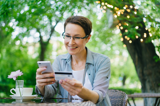 Радостная женщина платит кредитной картой, используя смартфон на открытом воздухе в уличном кафе
 - Фото, изображение