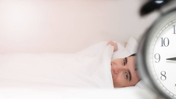 Будильник рядом с кроватью на фоне проснувшегося мужчины в постели
 - Фото, изображение
