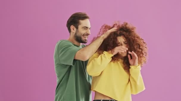 Handsome cheerful young couple smiling and playing with hair while having fun over pink background isolated - Video