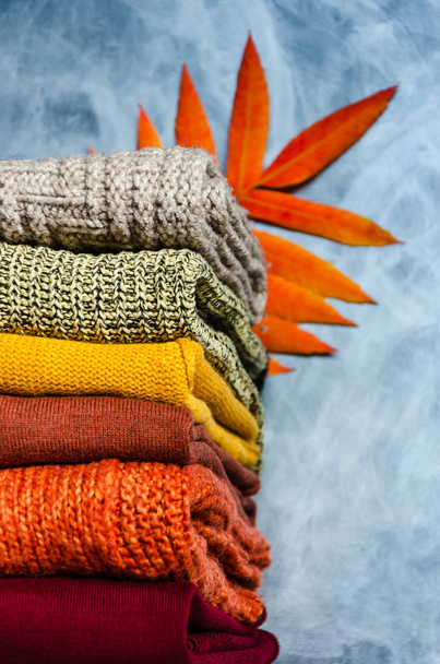 グレーの背景に暖かい色と明るい秋の葉で折り畳まれた暖かいニットの女性のセーターのスタック。クローズ アップ。スペースのコピー - 写真・画像