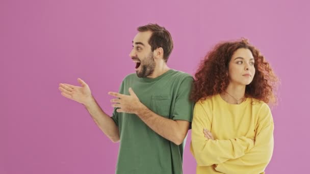 Handsome cheerful young bearded brunet man becoming surprised seeing something at the side and calling his girlfriend to show that. Upset young woman feeling annoyed because of boyfriend shaking her. Beautiful young couple over pink background - Footage, Video