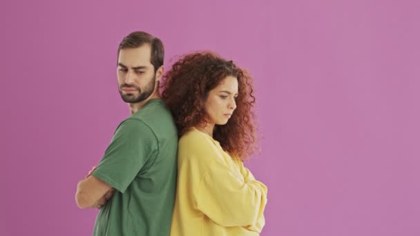 Upset young bearded brunet man turning around and hugging his girlfriend. Cute young redhead curly woman feeling upset at her boyfriend but forgiving and hugging him. Unhappy young couple staying back to back over pink background isolated - Кадры, видео