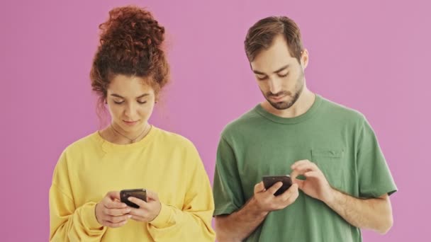 Attractive smiling young couple talking and laughing while using their smartphones over pink background isolated - Video