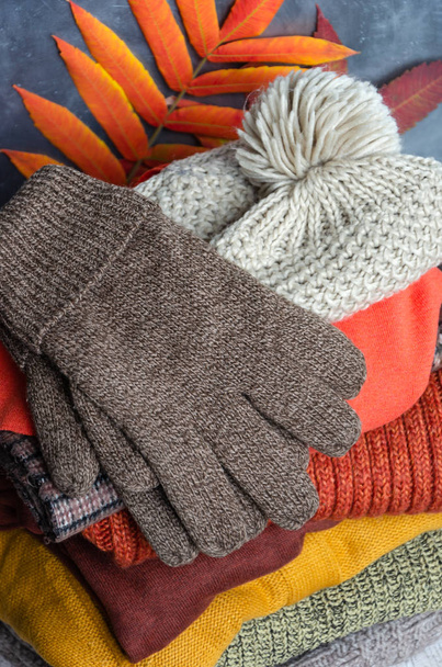 Montón de suéteres de punto cálido plegado para mujer, bufanda, sombrero con pompón, guantes en colores cálidos y hojas de otoño brillantes. Primer plano.
 - Foto, Imagen
