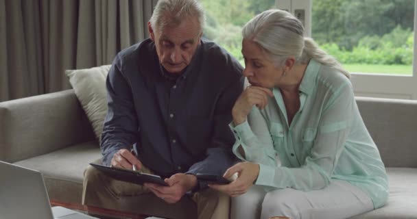 Front view of a worried senior Caucasian man and woman sitting on a couch and holding a smartphone, clipboard and using a laptop computer - Séquence, vidéo