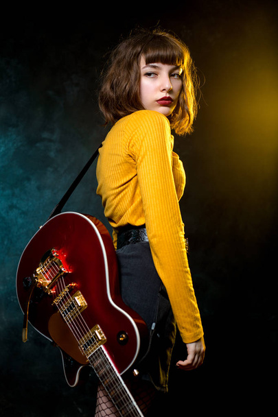 Πορτρέτο της όμορφης νεαρής χίππης γυναίκας με σγουρά μαλλιά με κόκκινη κιθάρα σε φώτα νέον. Ο ροκ μουσικός παίζει ηλεκτρική κιθάρα. Concept στυλ της δεκαετίας του ' 90. - Φωτογραφία, εικόνα