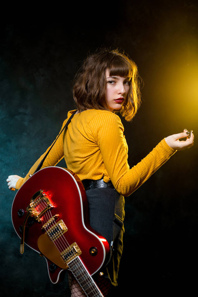 Portrait of beautiful young hipster woman with curly hair with red guitar in neon lights. Rock musician is playing electrical guitar. 90s style concept. - Zdjęcie, obraz