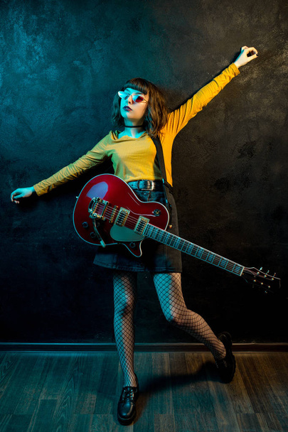 Fashion young hipster woman with curly hair with red guitar in neon lights. Rock musician is playing electrical guitar. 90s style concept. - Photo, Image