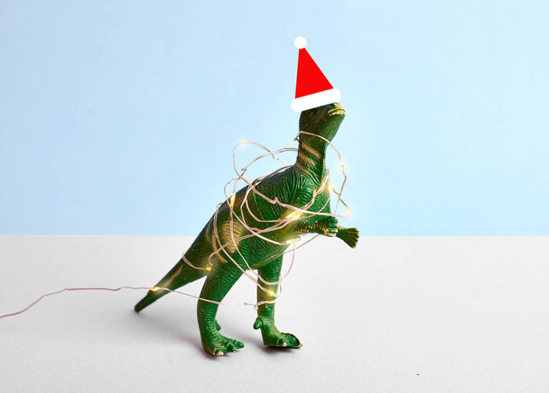 Zabawny zielony Tyrannosaur Rex w czerwonym Santa kapelusz i Boże Narodzenie światła. Niebieska koncepcja minimalistyczna - Zdjęcie, obraz