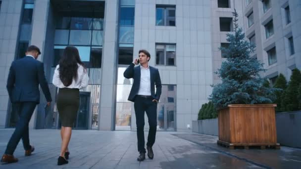 erfolgreicher Geschäftsmann, der auf den Straßen des Geschäftsviertels spaziert und mit dem Handy telefoniert. selbstbewusste attraktive kaukasische Mann im Anzug zu Fuß nach draußen in der Nähe von großen Bürogebäuden und mit Smartphone. - Filmmaterial, Video