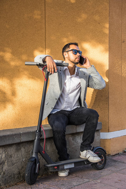 Latin felnőtt férfi napszemüveg, jól öltözött és elektromos robogó beszél a mobiltelefonján ül az utcán egy fekete vak a háttérben - Fotó, kép