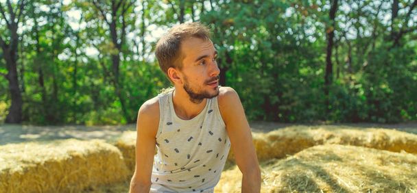 A young handsome man with beard of slim build sits in the hay and holds hay in his hands against the background of green trees in the park. - Fotó, kép