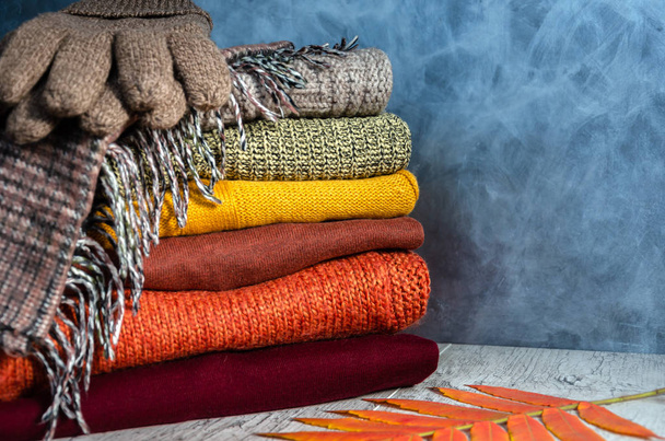 Pilha de camisolas femininas de malha quente dobrada em cores quentes, cachecol xadrez, luvas de malha e folhas de outono no fundo cinza. Fecha a porta. Espaço de cópia
 - Foto, Imagem