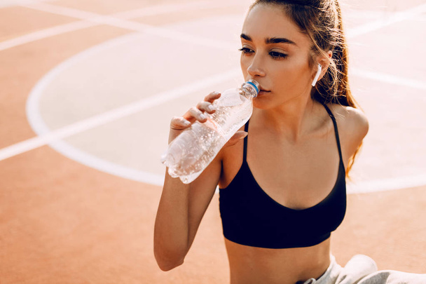 Sporty girl, in top and sweatpants, drinking water from a bottle, sitting on the field after a workout - Zdjęcie, obraz