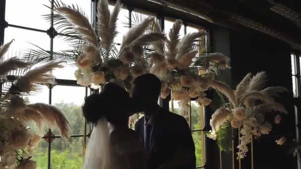 La novia y el novio se besan en el fondo de una gran ventana y un arco de flores
. - Imágenes, Vídeo