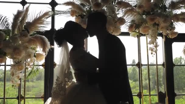 Наречений і наречений цілуються на тлі великого вікна і арки квітів
. - Кадри, відео