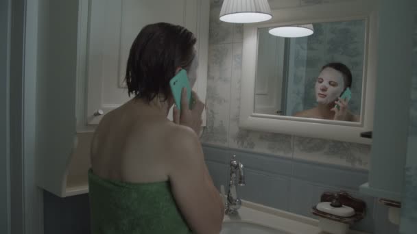 Slow motion of brunette female in towel in cosmetic mask on her face with smartphone after shower  - Filmmaterial, Video
