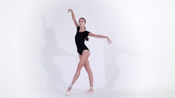 Ballerina in black suit practicing in ballet in light studio. Young girl isolated on white wall dancing classic dance in pointe shoes. Slow motion.  - Záběry, video