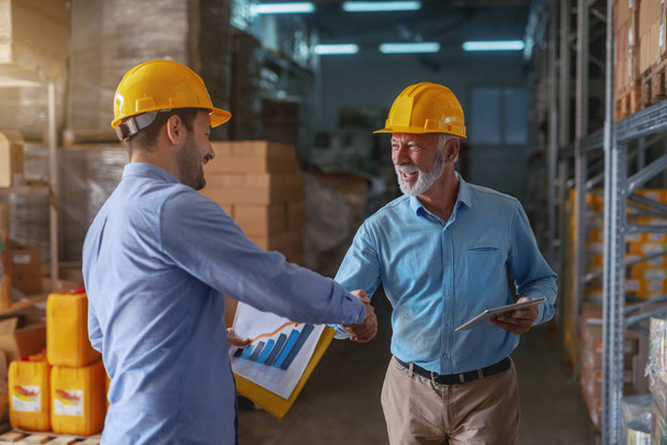 Supervisor congratulating for good results in sale. Younger man holding folder with data while older one holding tablet. Both having yellow helmets on heads. Warehouse interior. - Foto, imagen