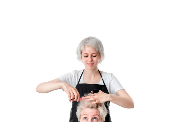Αρκετά νέος κομμωτής λευκό μπλουζάκι και μαύρη ποδιά κόβοντας τα μαλλιά σε μια ξανθιά με κοντό κούρεμα. απομονώνονται σε λευκό φόντο. Ο πελάτης αν είναι λίγο νευρικός για το αποτέλεσμα. - Φωτογραφία, εικόνα