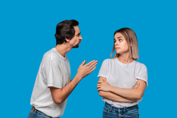 Brunet bearded man with mustaches in white t-shirt and blue jeans asking his girlfriend for forgiveness isolated over blue background. Concept of guilt. - Фото, изображение