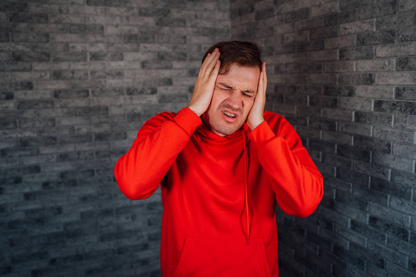 A young man in a red hoodie grabs her head with hands in pain on a brick background. A male has a headache. - Foto, Imagen