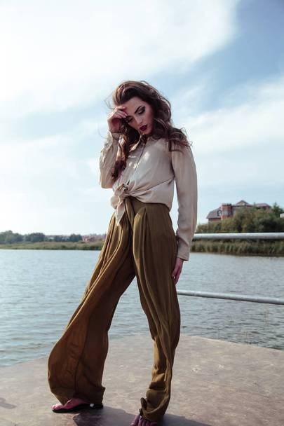 Young fashionable woman in stylish clothes posing near river side in a summer evening. Fashion model - Zdjęcie, obraz