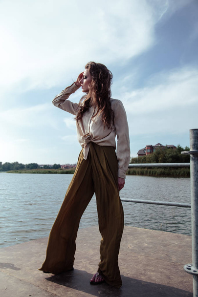 Young fashionable woman in stylish clothes posing near river side in a summer evening. Fashion model - Fotoğraf, Görsel