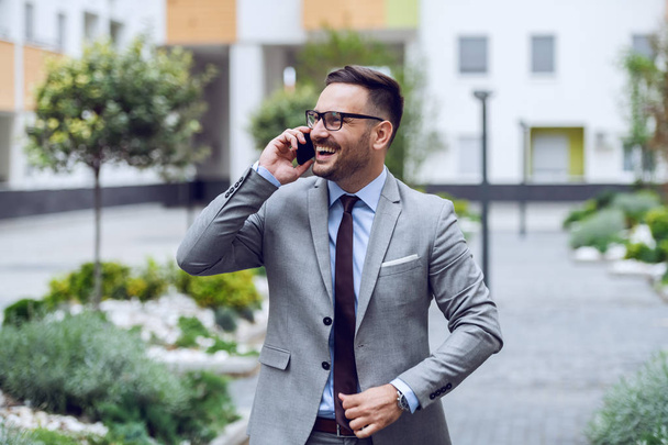 Smiling caucasian businessman dressed in formal wear and with eyeglasses talking on the smart phone. Business center exterior. - Foto, Imagem