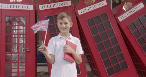 Lassú mozgás portréja Aranyos fiú integetett brit zászló áll a szabadban egyedül mosolyogva nézi kamera. A háttérben angol piros telefon fülkék. Utazás koncepció. - Felvétel, videó