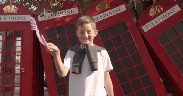 Slow motion portrait of cute boy waving British flag standing outdoors alone smiling looking at camera. On the background English red telephone booths. Travelling concept. - Footage, Video