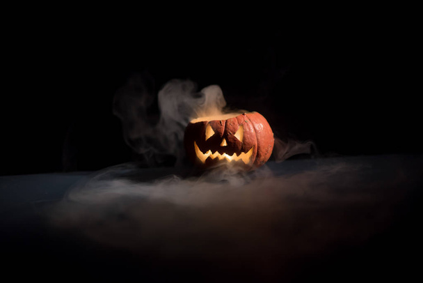 Halloween, orange pumpkin with a scary luminous face on a dark background. Thick gray smoke comes out and spreads across the black table. A close-up of a flashlight on the eve of all the saints - 写真・画像