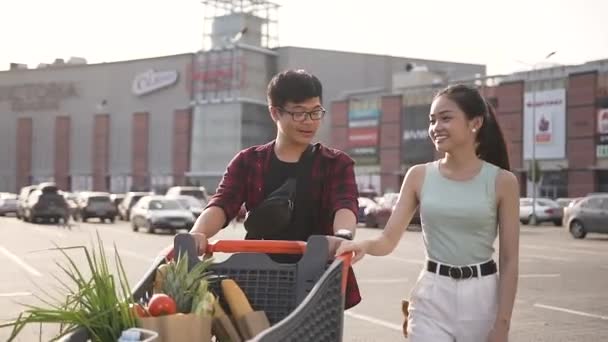 Happy attractive asian couple being pushed in front of them a cart full of various products - Filmati, video