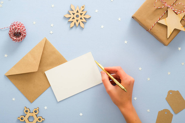 Female hand writing letter to Santa Claus over blue background with Christmas decorations, confetti stars, wooden snowflakes, gift box wrapping kraft paper. Christmas, New Year, winter holiday concept - Photo, image