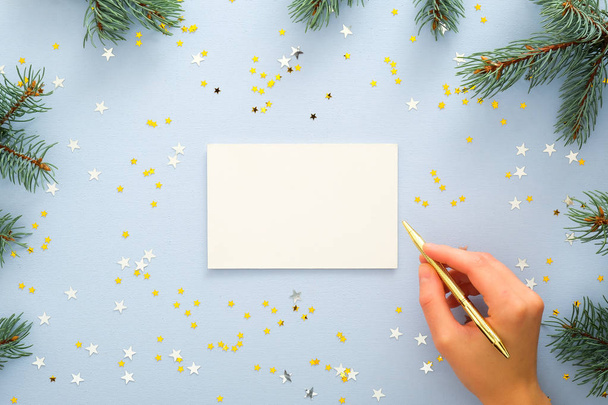 Top view of woman hands writing on a blank paper greeting or invitation card over blue background with fir tree branches and glitter confetti stars. Christmas, New Year, winter holidays concept - Photo, image