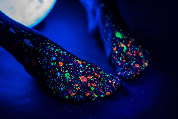Barefoot female feet in a spray of paint glowing in the dark. Toes in fluorescent powder. Body art glowing in ultraviolet light, like a blue night sky with stars. Close up. - Photo, Image