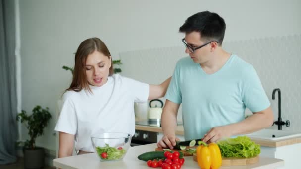 Man cooking salad cutting cucumber feeding girlfriend kissing in kitchen at home - Metraje, vídeo