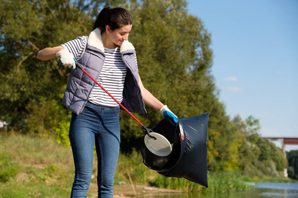 Volunteer woman picking up plastic garbage on coast of the river. Cleaning environment concept - Foto, afbeelding